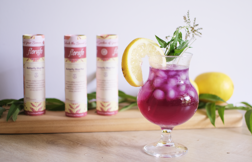 A refreshing summer mocktail drink of florafo Butterfly Blue Pea Lemonade, with ice cubes, a slice of lemon and rosemary. In the background are three florafo butterfly blue pea paper tubes and two lemons on a wooden serving board.