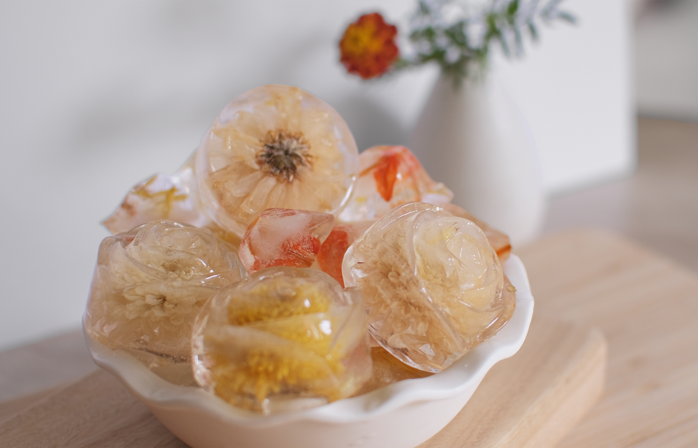 florafo floral ice cubes containing florafo mixed flower tea ball flowers, sitting in a white ceramic bowl on a wooden serving board, perfect for adding to your summer drinks for a refreshing cool and tasty taste.