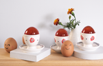 Three Easter jelly eggs made from florafo Rose & Osmanthus tea balls, each sitting in an egg cup on a wooden serving board. Two egg shells and a marigold cutting complement the image.